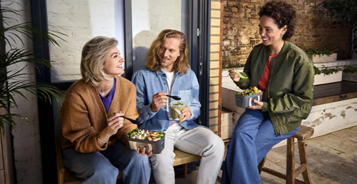 Drei Personen sitzen draußen auf einer Bank. Sie essen Salate und Suppen to go aus Mehrweg-Pfandbehältern.