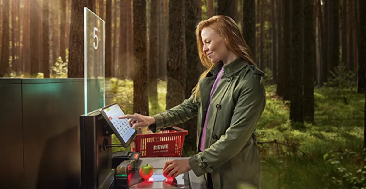 Eine moderne Selbstbedienungskasse steht mitten im Wald. Eine Person nimmt Produkte aus einem roten REWE Einkaufskorb und scannt sie ein.