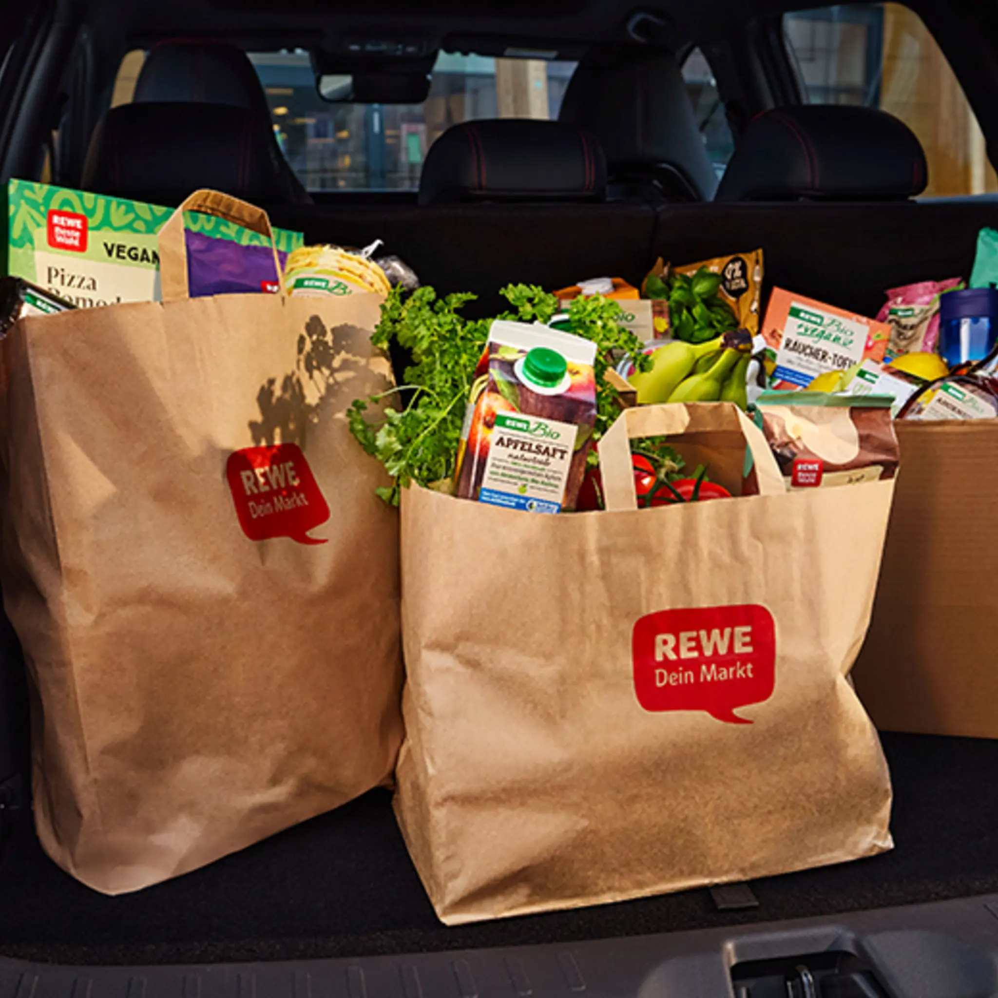 Ein Karton und zwei REWE Papiertüten gefüllt mit Lebensmitteln im Kofferraum eines Autos