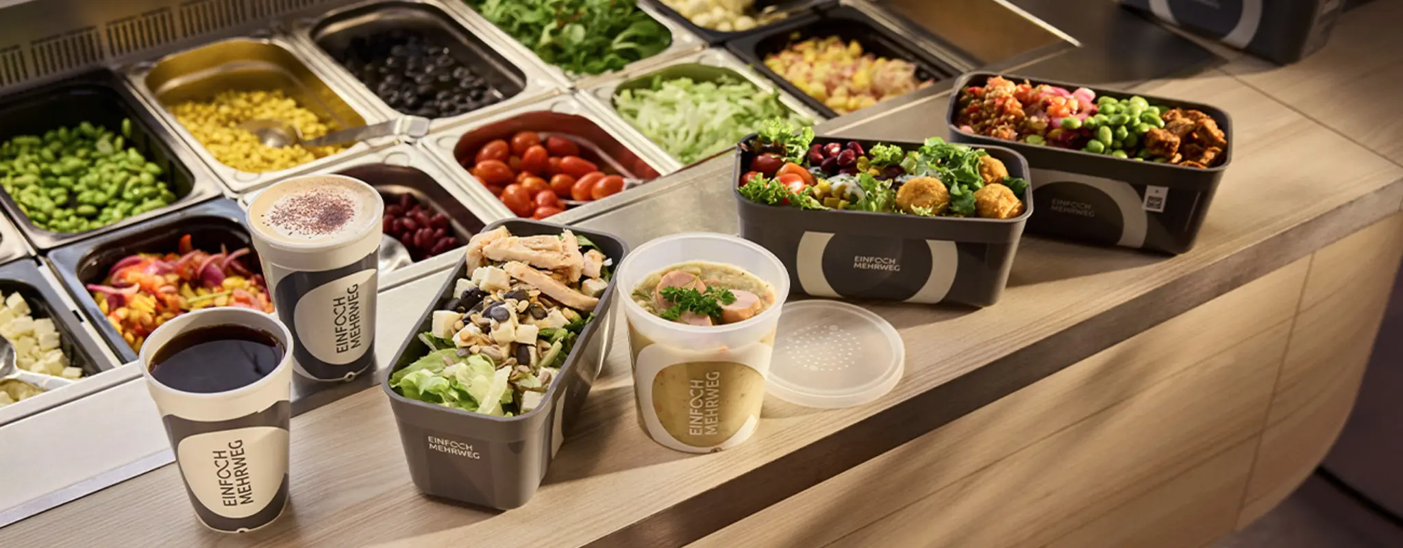 Speisen und Getränke in Mehrwegverpackungen auf dem Tresen vor einem Salatbuffet   