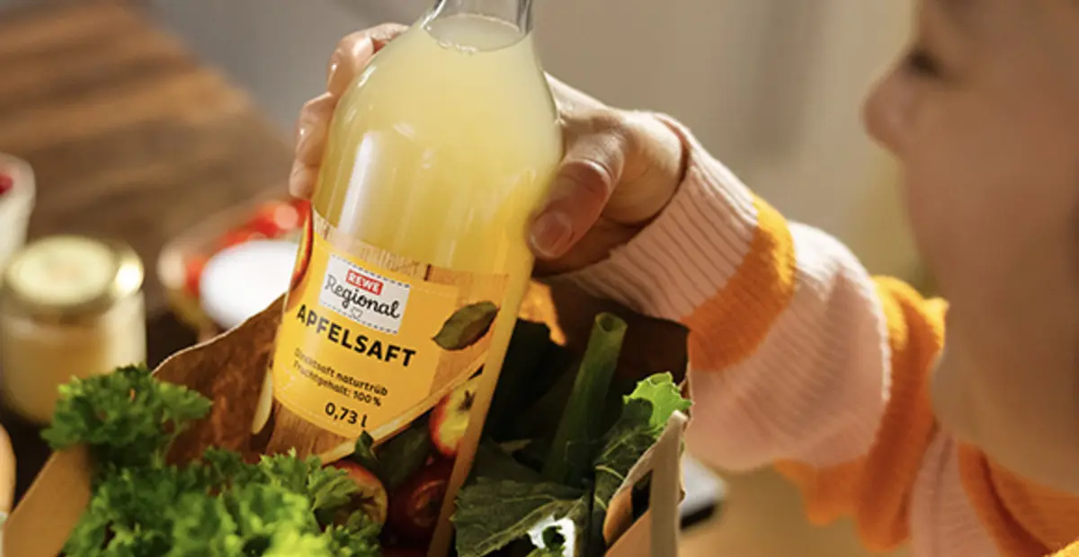 Eine Frau holt eine Flasche REWE Regional Apfelsaft auf einer REWE Papiertragetasche voll frischem Obst und Gemüse. 