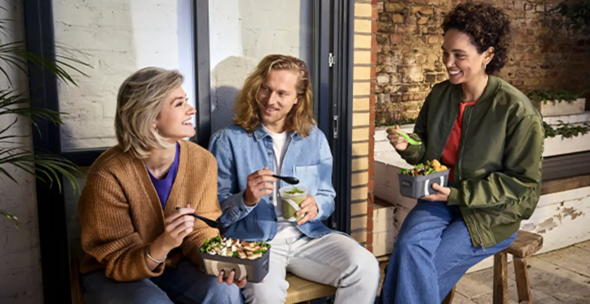 Drei Personen sitzen draußen auf einer Bank. Sie essen Salate und Suppen to go aus Mehrweg-Pfandbehältern.