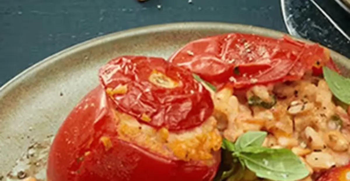 Ein Teller mit gefüllten Tomaten steht auf einem gedeckten Tisch. Im Hintergrund steht eine Schale mit Pinienkernen und ein Stängel Basilikum liegt neben dem Teller.