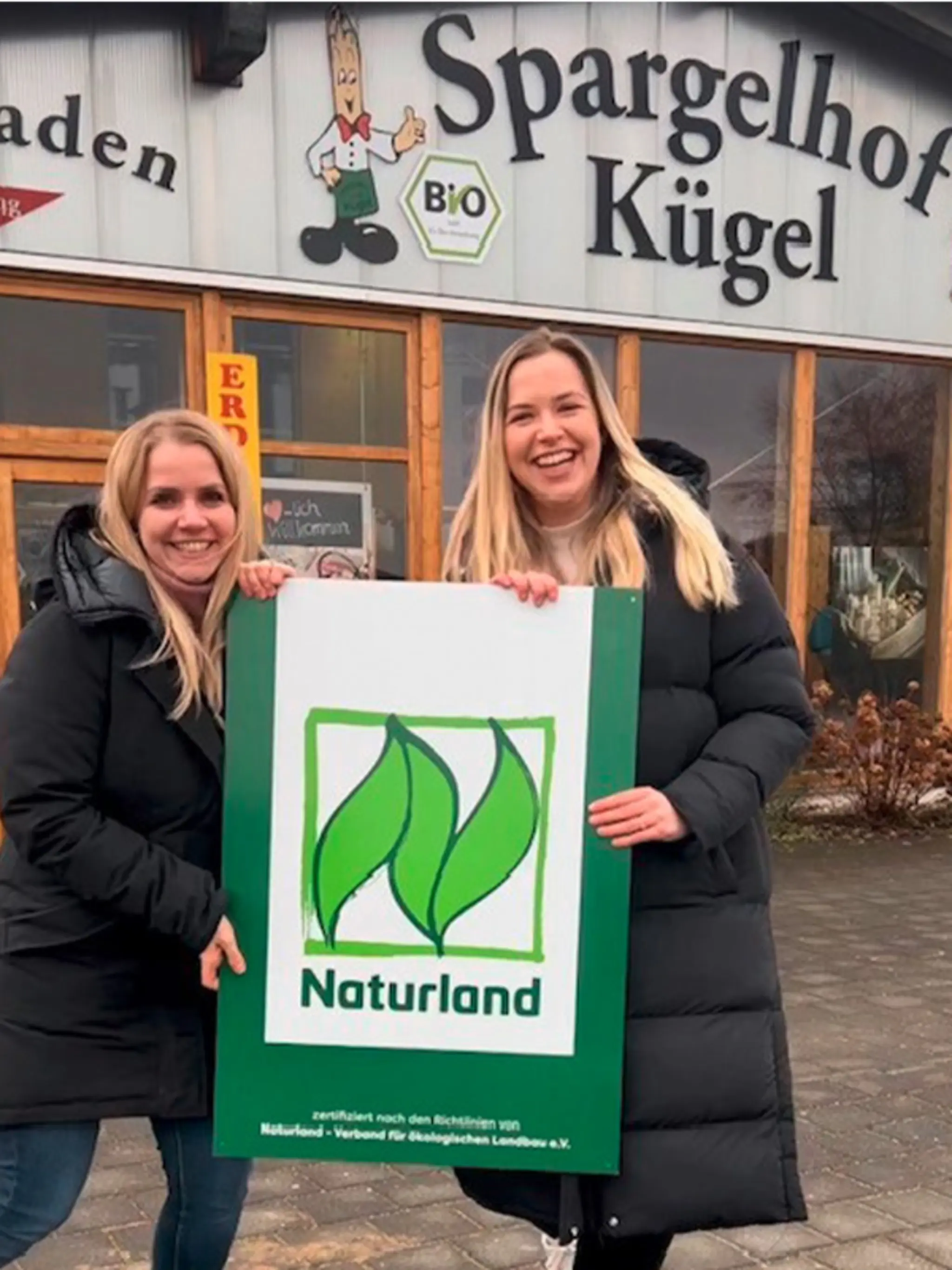 Zwei der Töchter der Familie Kügel halten zwischen sich ein großes Schild mit dem Naturland Logo. Als REWE Wegbereiter haben sie sich im Öko-Verband angemeldet.