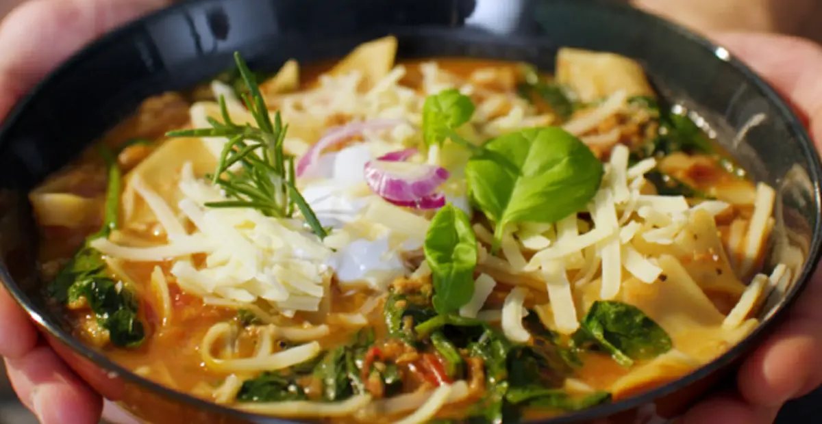 Eine Schüssel Tomaten-Suppe mit darin gekochten Lasagne-Platten, geriebenem Käse, frischen Basilikum und Rosmarin. 
