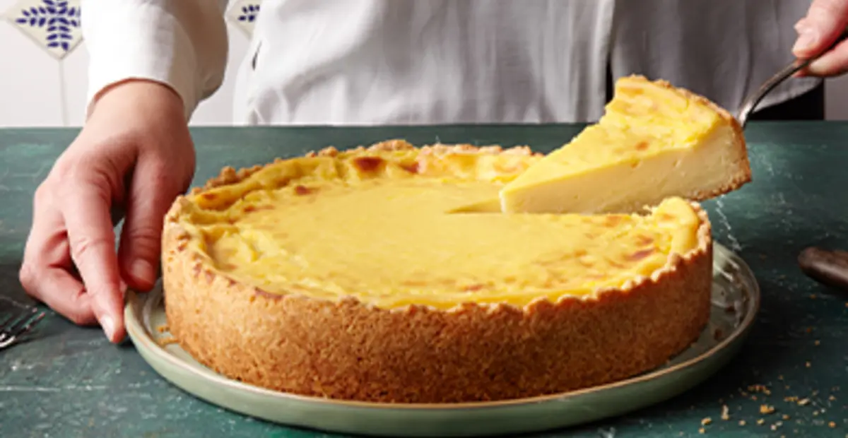 Ein angeschnittener veganer Käsekuchen. Eine Hand nimmt mit einem Tortenheber das erste Stück heraus.