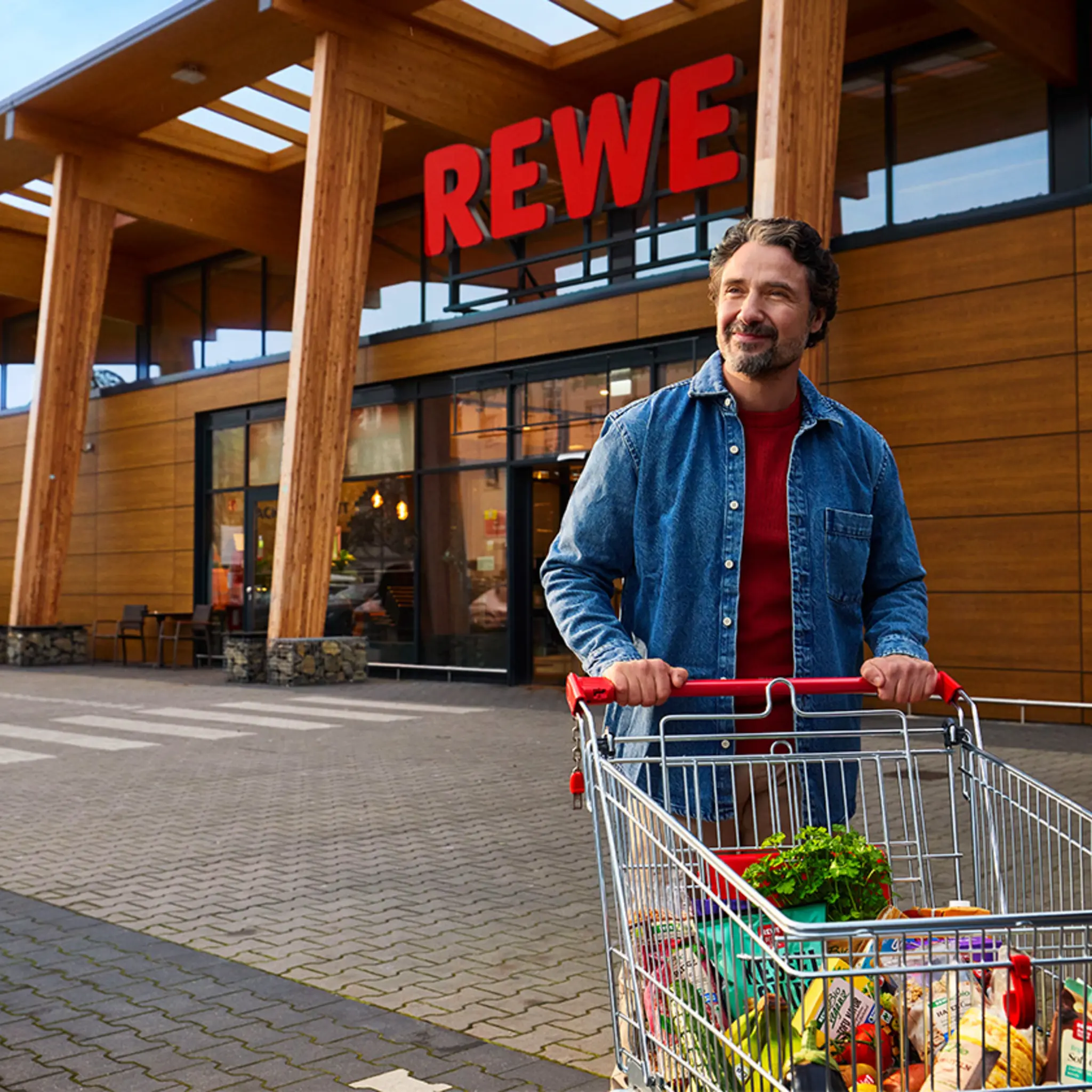 Ein Kunde kommt aus einem nachhaltigeren REWE Markt mit Holz- und Glasfassade.
