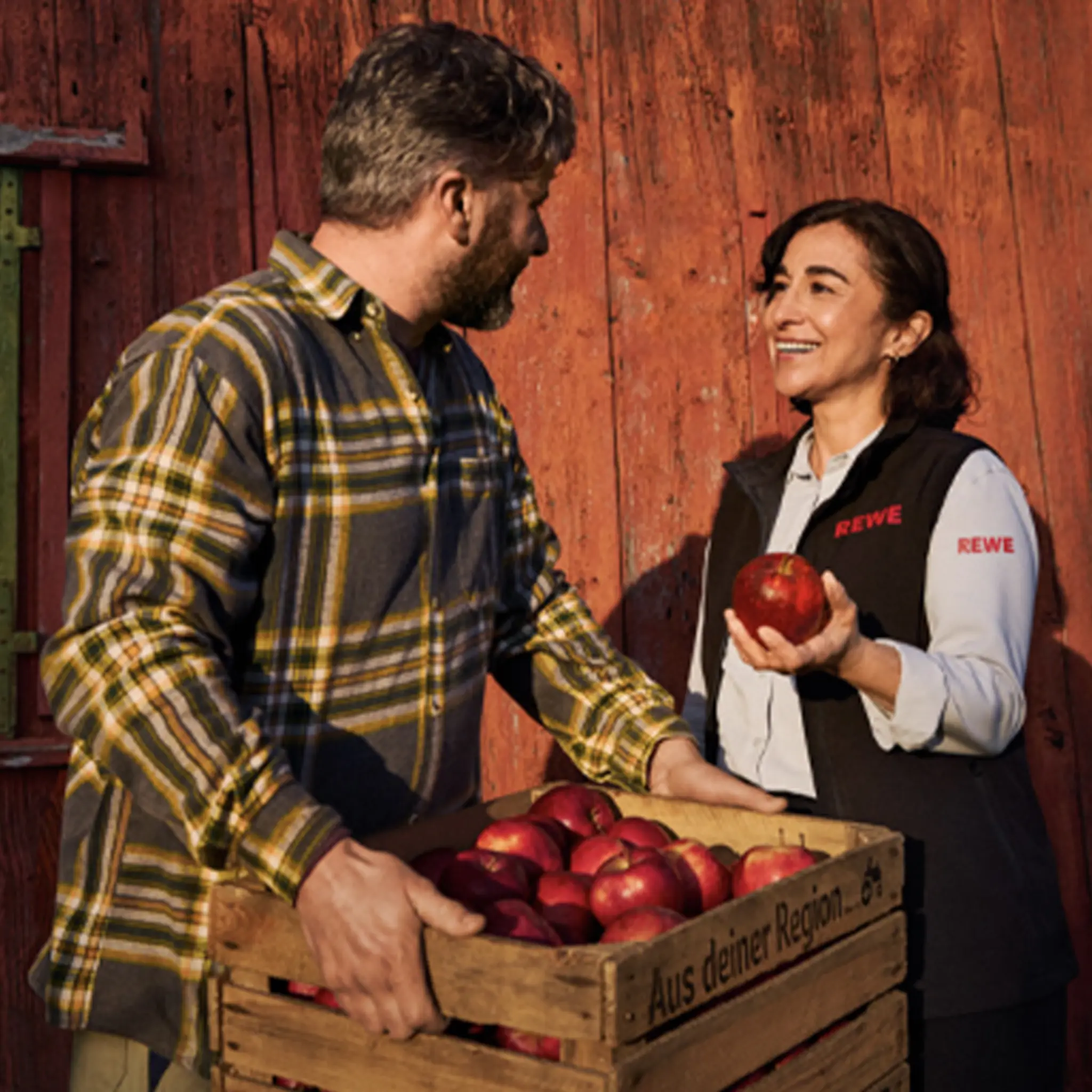 Eine REWE Mitarbeiterin im Gespräch mit einem regionalen Landwirt. Die Darstellung vermittelt: Sie weiß, woher die regionalen Äpfel in ihrem REWE Markt kommen und geht partnerschaftlich mit den Erzeuger:innen um.