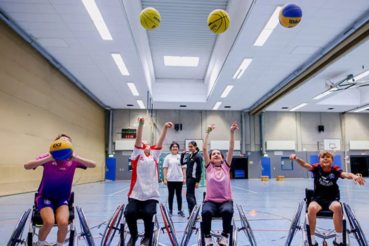 Mehrere Kinder im Rollstuhl werfen auf einen Basketballkorb. 