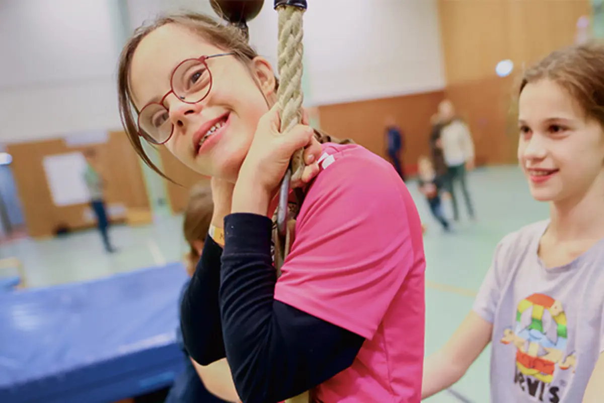 Ein Kind mit Behinderung schwingt in einer Turnhalle an einem Tau. Ein zweites Kind hilft ihm. 