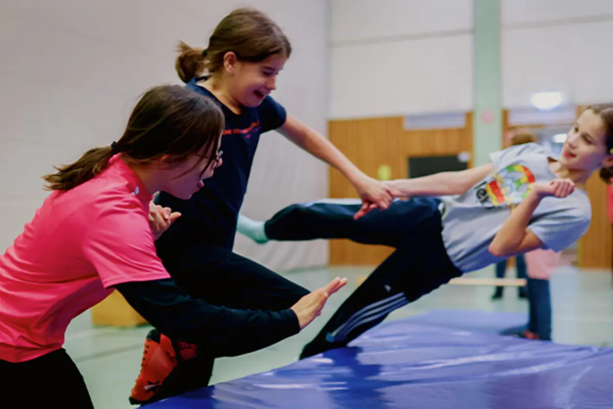 In einer Sporthalle. Drei Kinder mit und ohne Behinderung lassen sich gemeinsam auf eine dicke Gymnastikmatte fallen. 