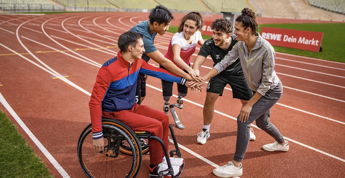 Menschen mit und ohne Behinderung legen ihre Hände aufeinander, um sich auf einer Tartanbahn auf ein sportliches Event einzustimmen. 