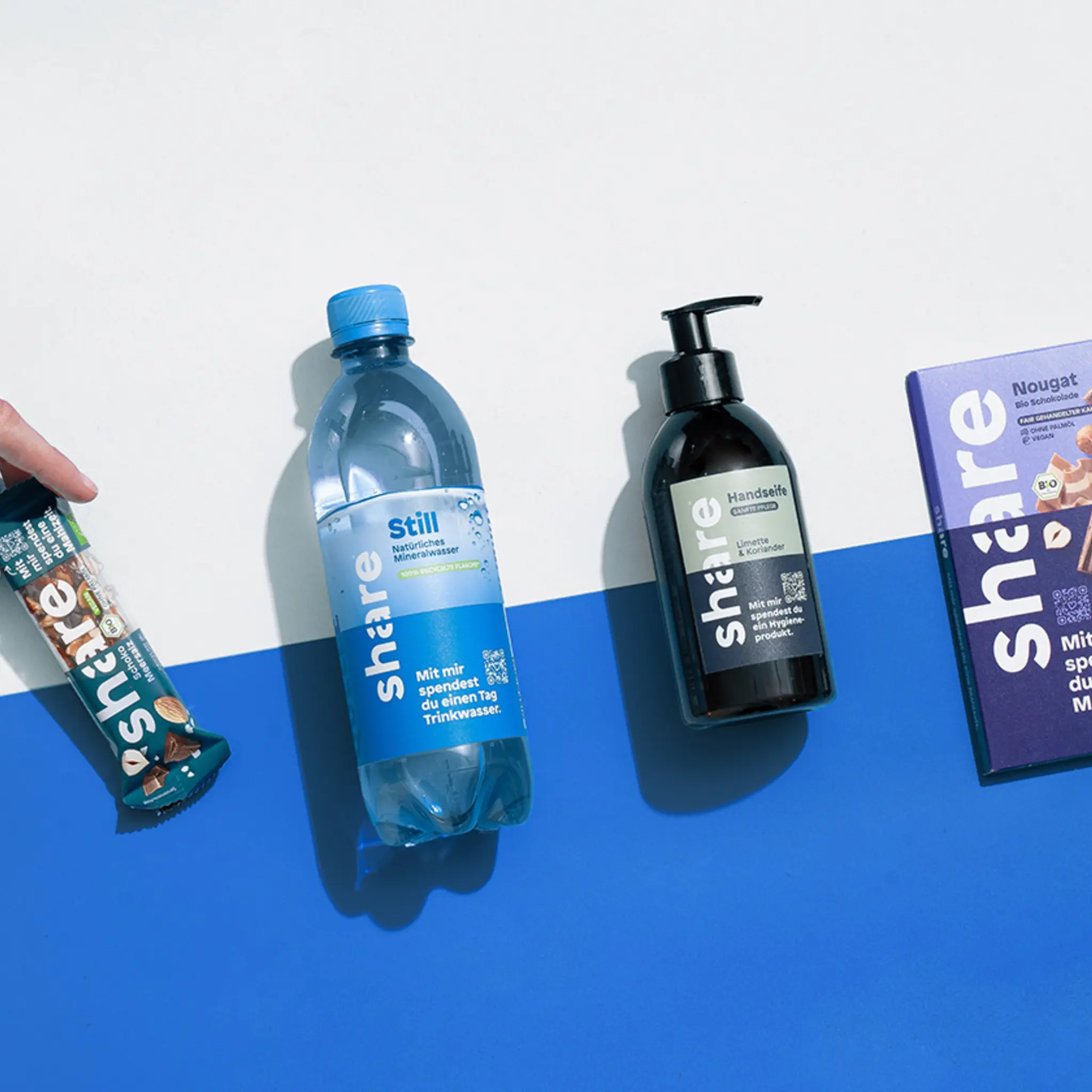 Eine Hand greift nach einem share Müsliriegel, eine Flasche share stillem Wasser, einer Flasche share Seife und einer Tafel share Schokolade. 