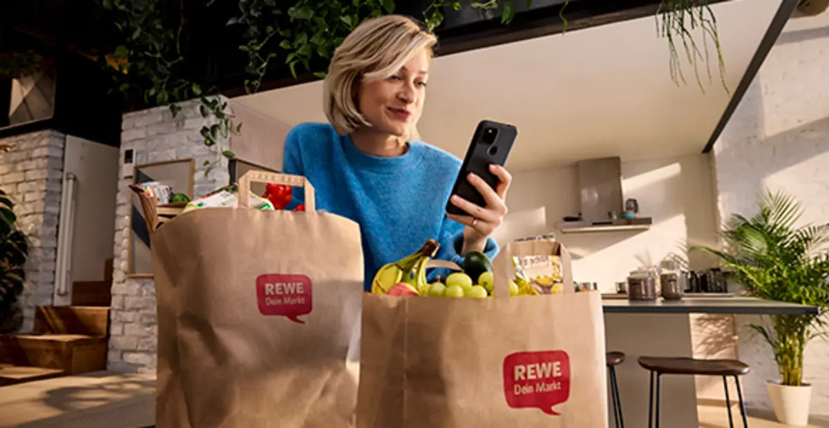 Eine Person steht mit vollen REWE Einkaufstüten in der Küche. Sie schaut aufs Handy.