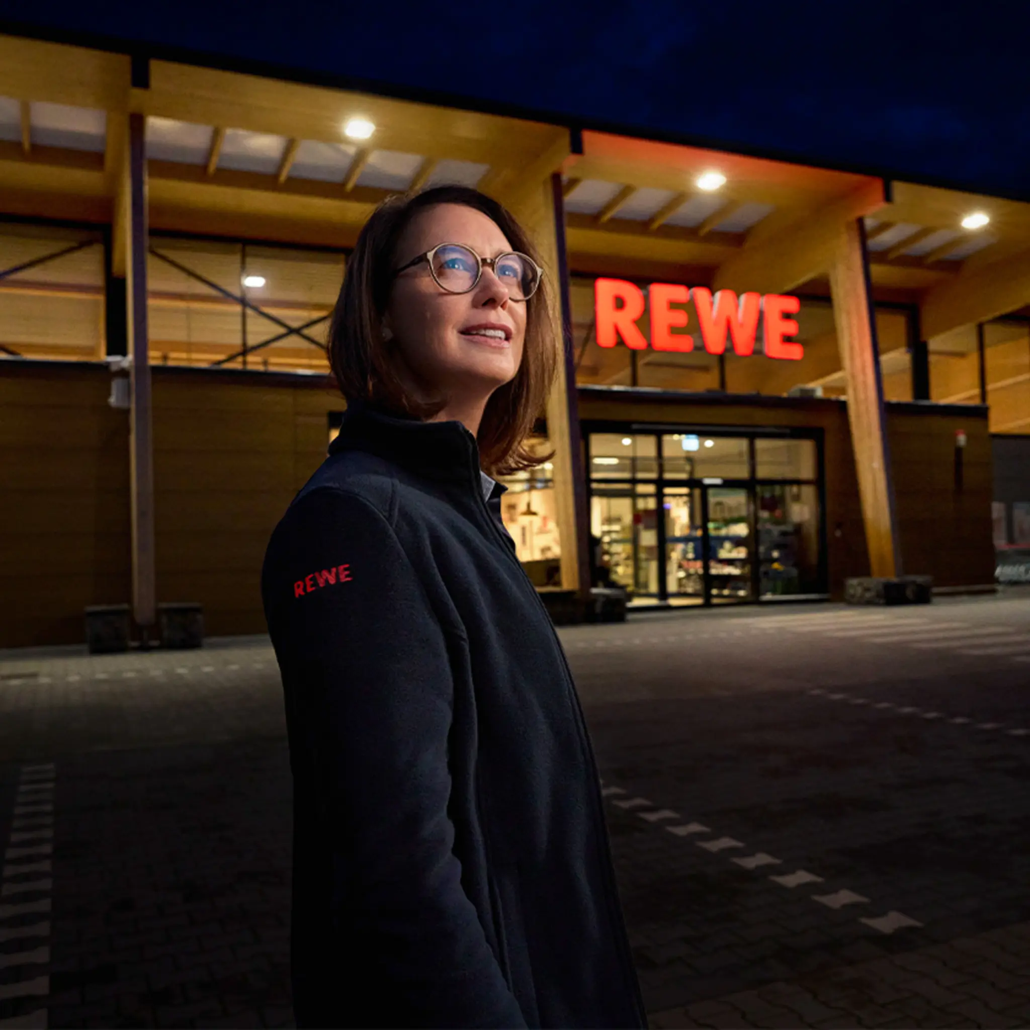 Ein beleuchteter REWE Markt am Abend. Davor steht eine Person, die bei REWE arbeitet.