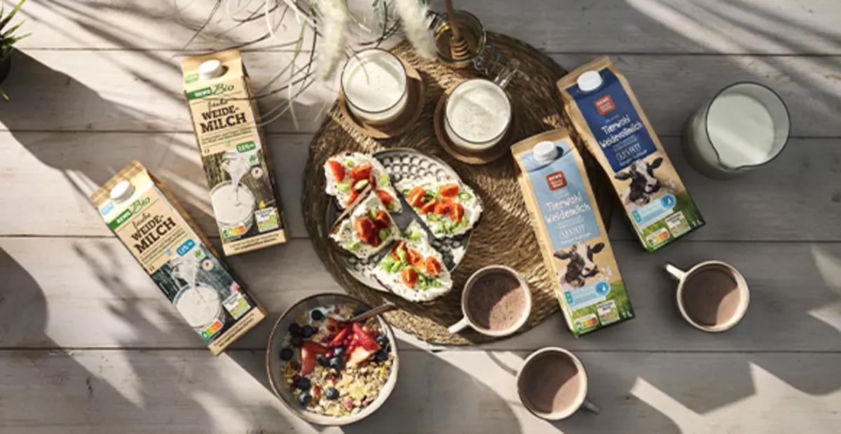 Verschiedene REWE Produkte aus höheren Haltungsstufen liegen auf einem Tisch. Dazwischen stehen Gläser voll Milch und eine Müslischüssel.