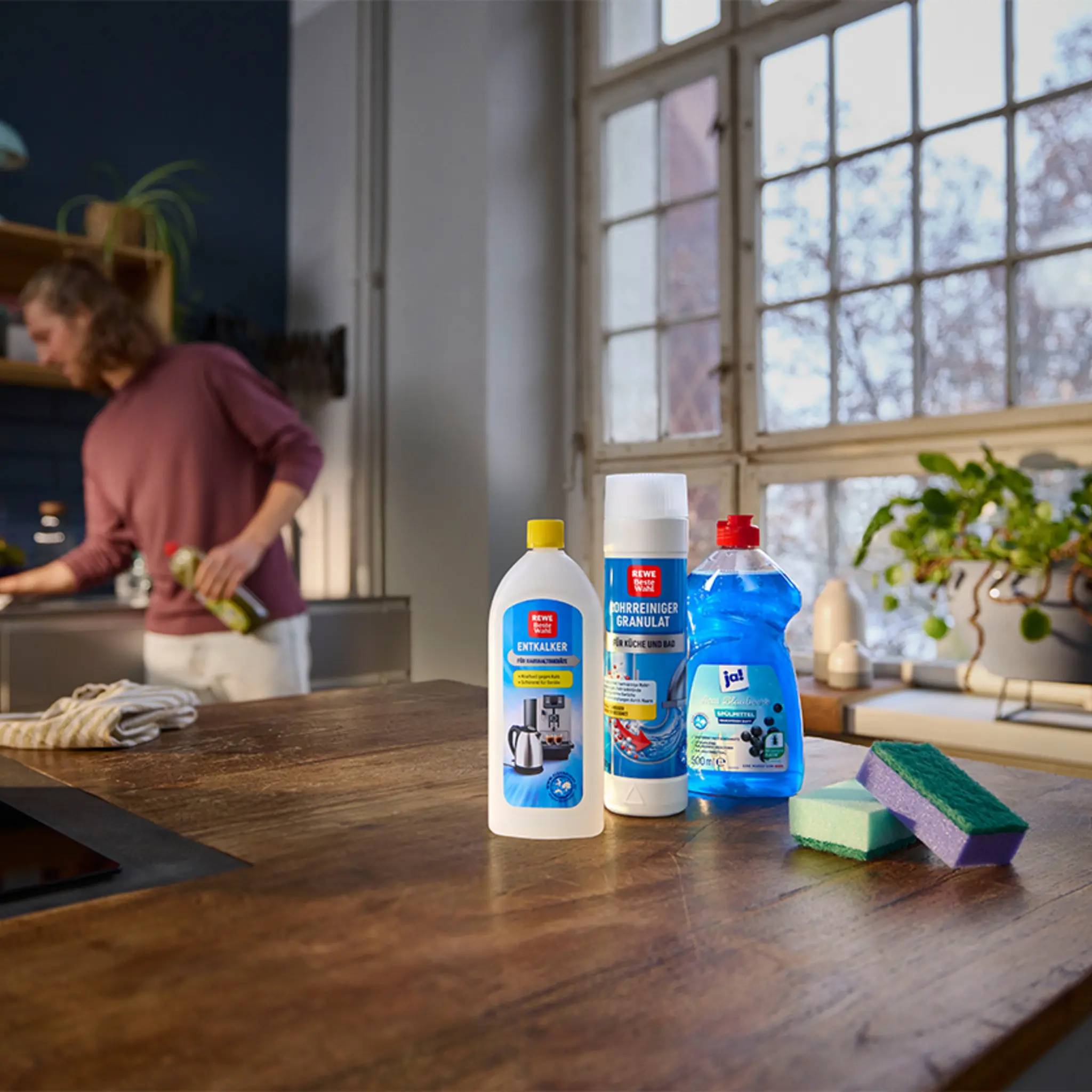 Eine Person putzt die Küche. Auf dem Tresen stehen eine Flasche REWE Beste Wahl Entkalker, eine Flasche REWE Beste Wahl Rohrreiniger Granulat und eine Flasche ja! Spülmittel.
