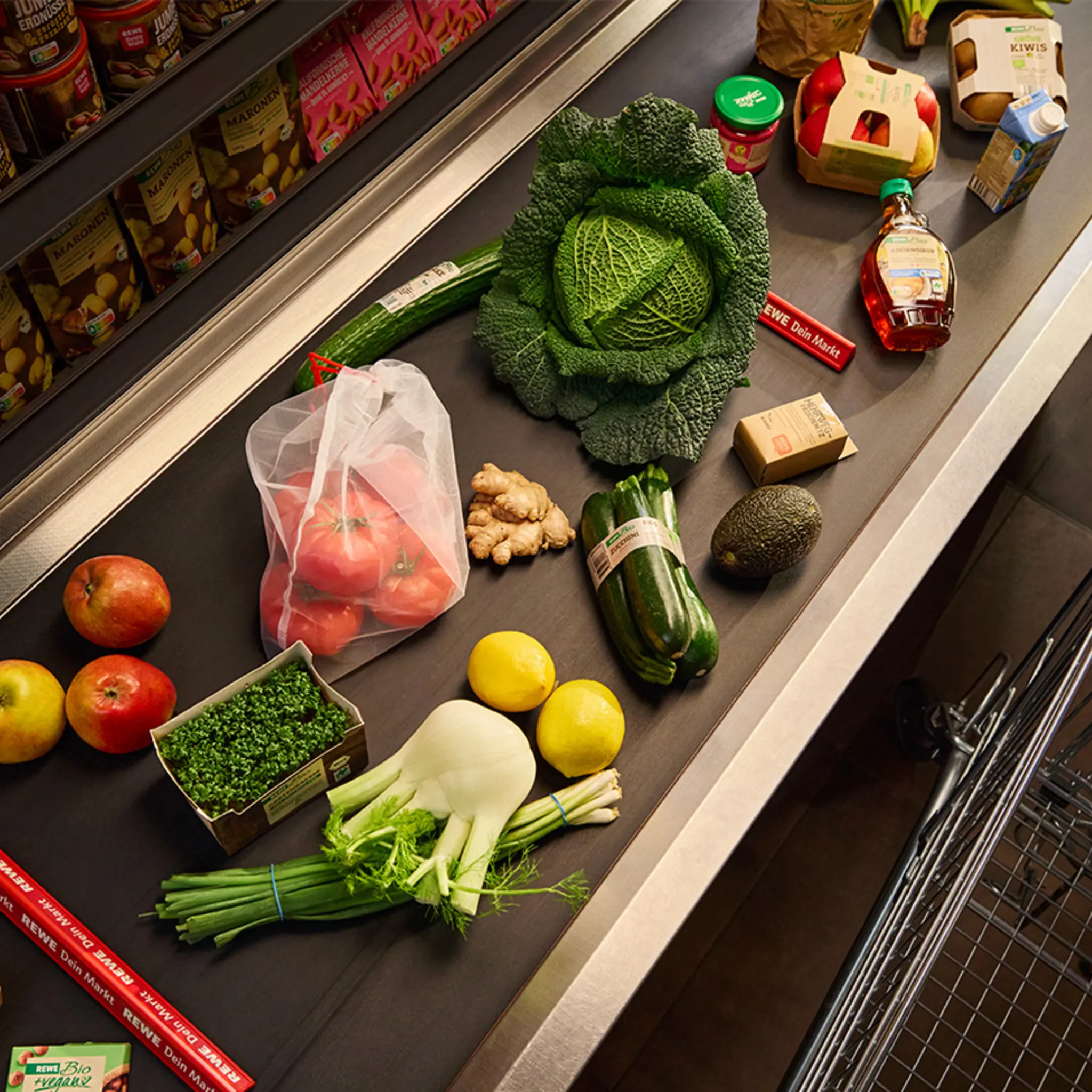 Viele unterschiedliche Lebensmittel liegen auf einem Kassenband. Einige von ihnen sind unverpackt. Bei anderen wurde die Verpackung verbessert. Zum Beispiel werden drei REWE Bio Zucchini von einer Banderole zusammengehalten.