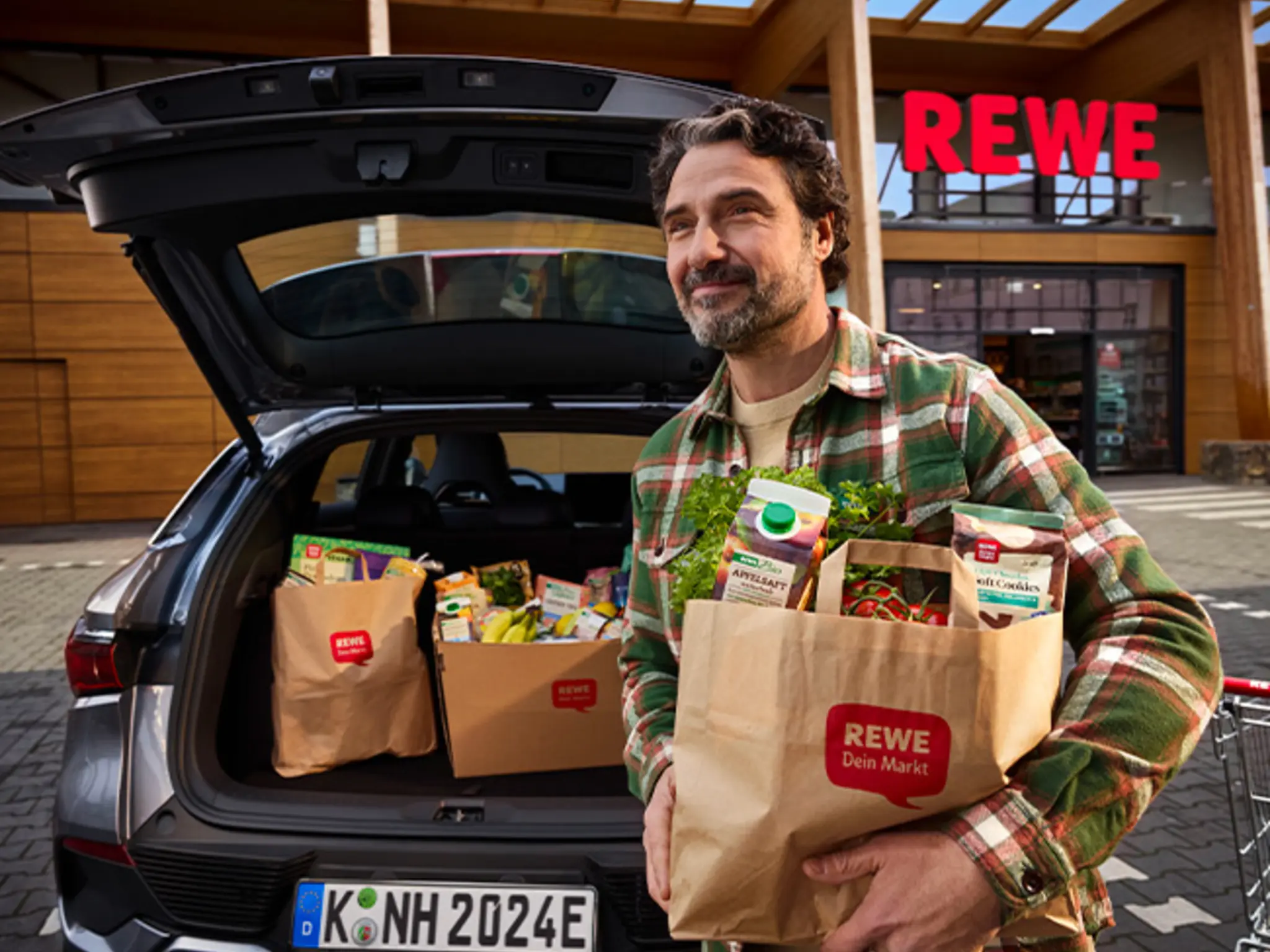 Eine Person lädt REWE Tüten und Transportboxen aus zertifiziertem Material in den Kofferraum eines Autos.