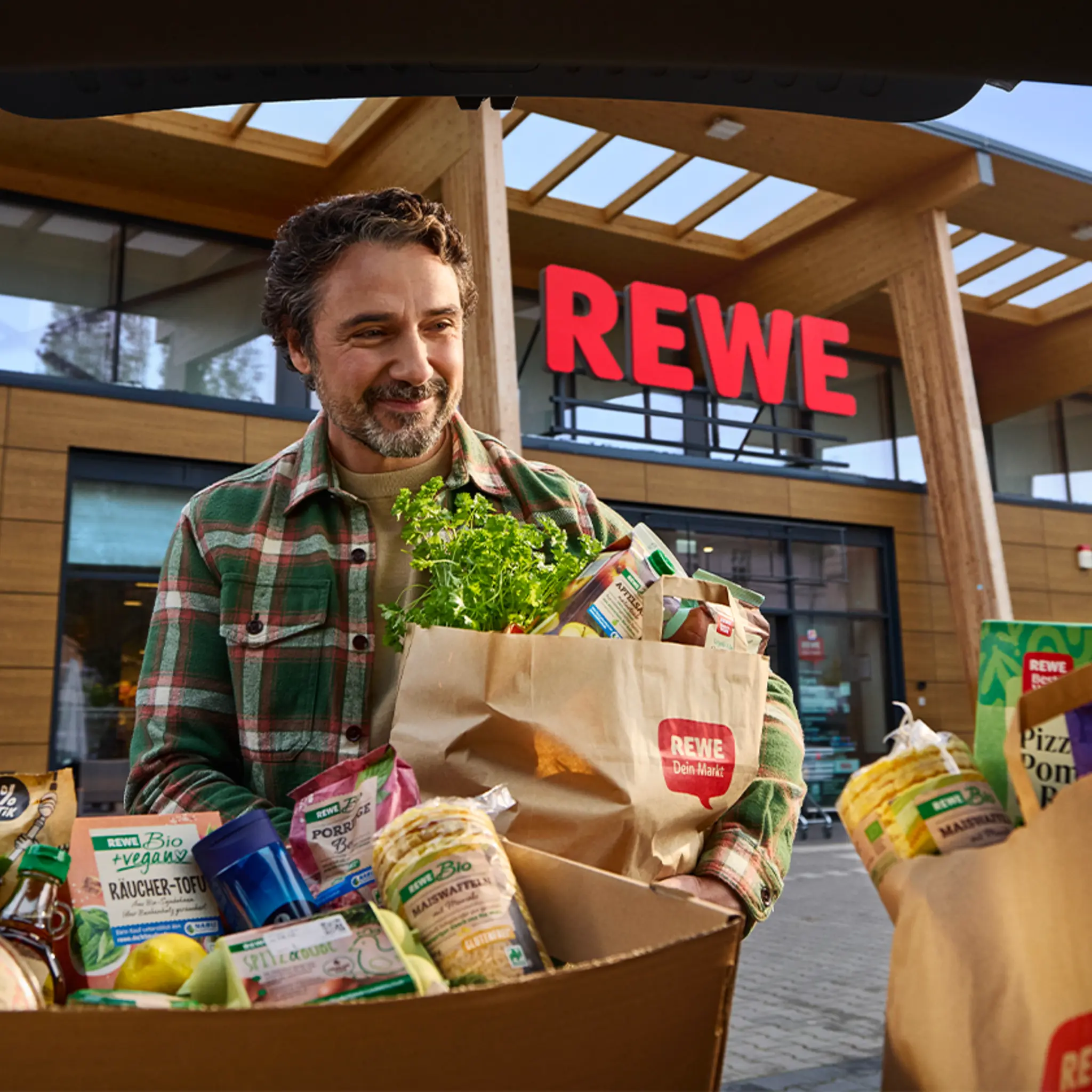 Vor einem REWE Markt. Eine Person hat den Einkauf in verschiedene REWE Tüten und Boxen verpackt.