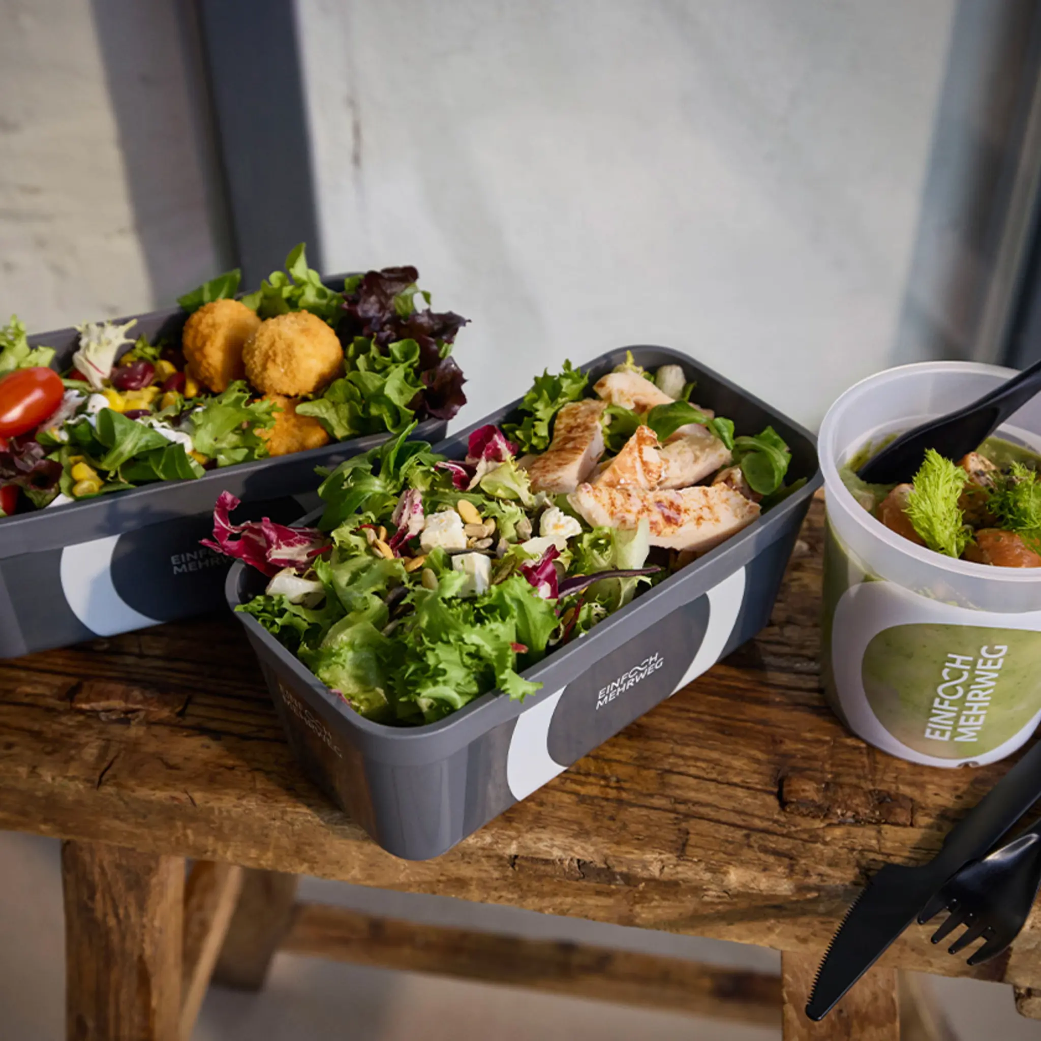 Drei Mehrweg-Pfandbehälter mit frischen Salat und Suppe stehen auf einem kleinen Holztisch. 