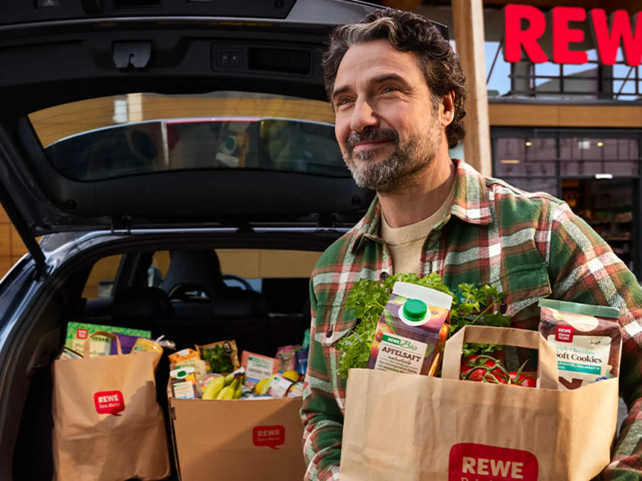 Auf dem Parkplatz eines REWE Markts. Eine Person lädt mehrere volle Papier-Tragetaschen in den Kofferraum.
