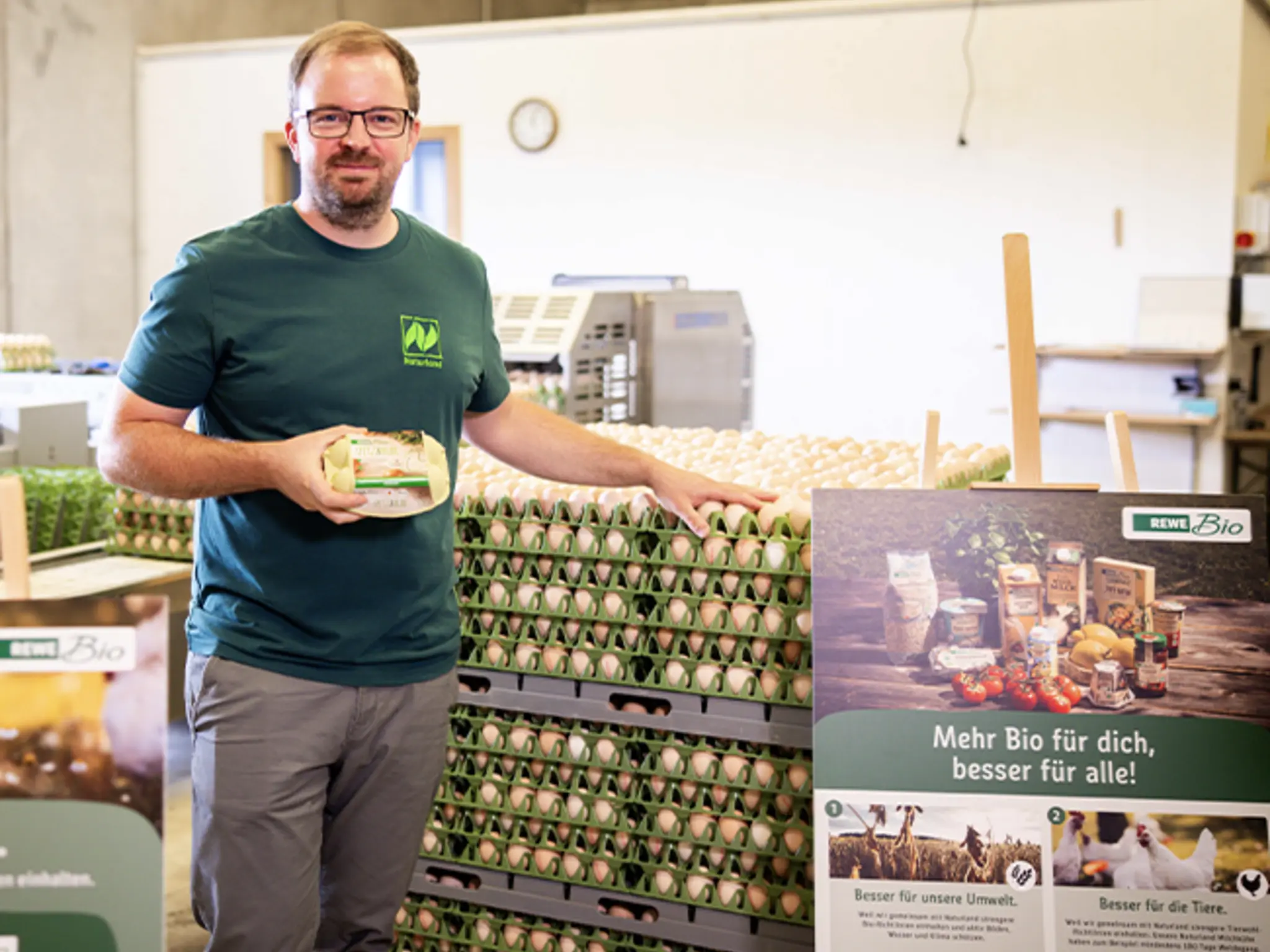 Christoph Möller hält eine Packung REWE Spitz & Bube Eier in der Hand. Ein großes Plakat von REWE Bio trägt die Aufschrift „Besser für unsere Tiere“.