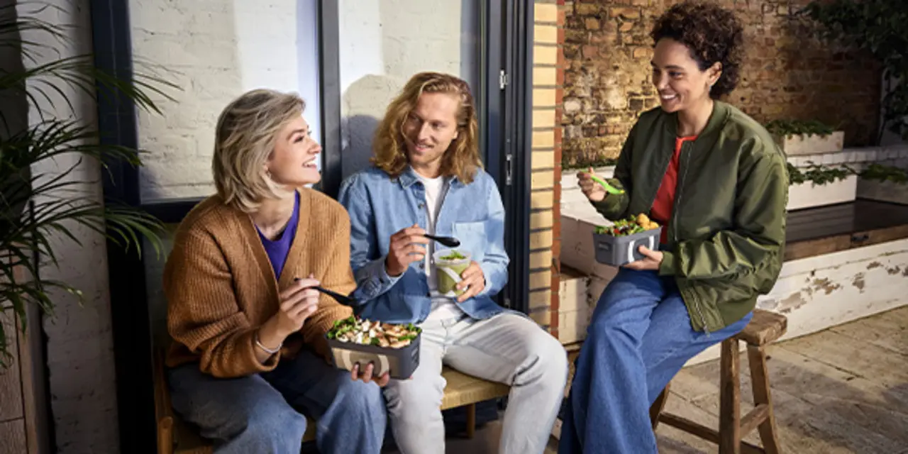 Drei Personen sitzen draußen auf einer Bank. Sie essen Salate und Suppen to go aus Mehrweg-Pfandbehältern.
