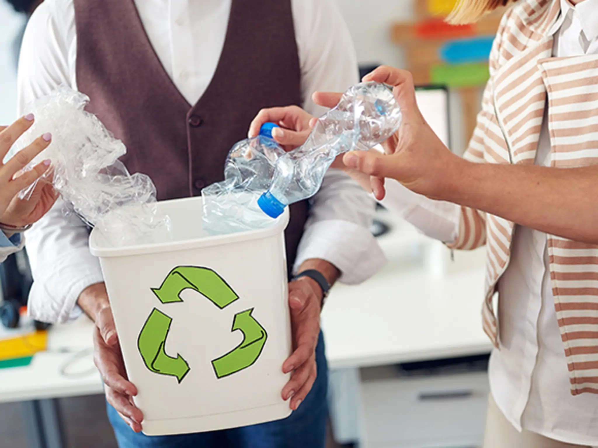 Ein Mann hält einen weißen Recyclingbehälter in den Händen, in den weitere Personen Plastikflaschen werfen. 