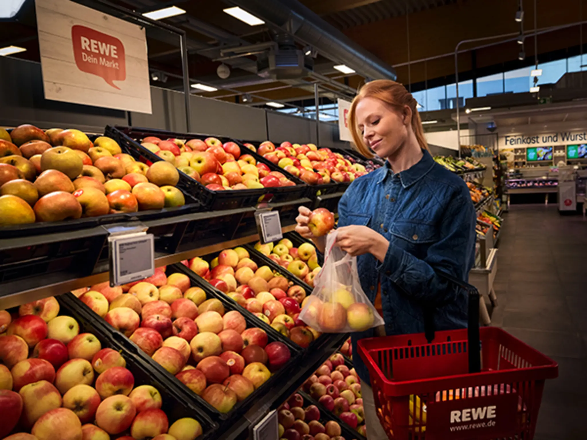 Eine lächelnde Frau im Jeanshemd legt in der REWE-Obstabteilung Äpfel in ein wiederverwendbares Obstnetz. 