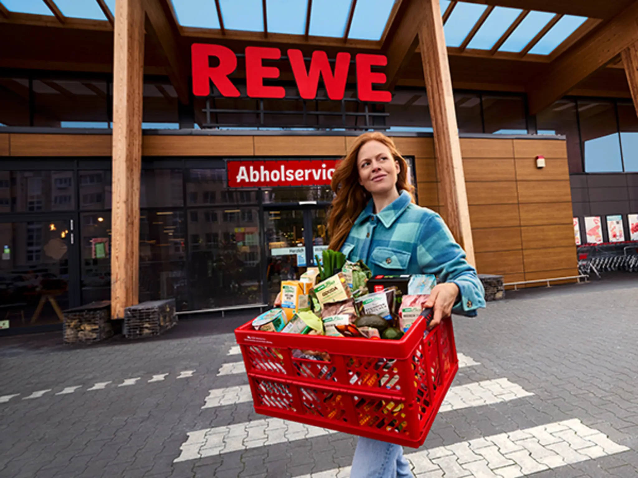 Eine Frau in einer blau-karierten Jacke steht mit einer Einkaufkiste voller REWE Bio-Produkten vor dem Abholservice einer REWE-Filiale. 