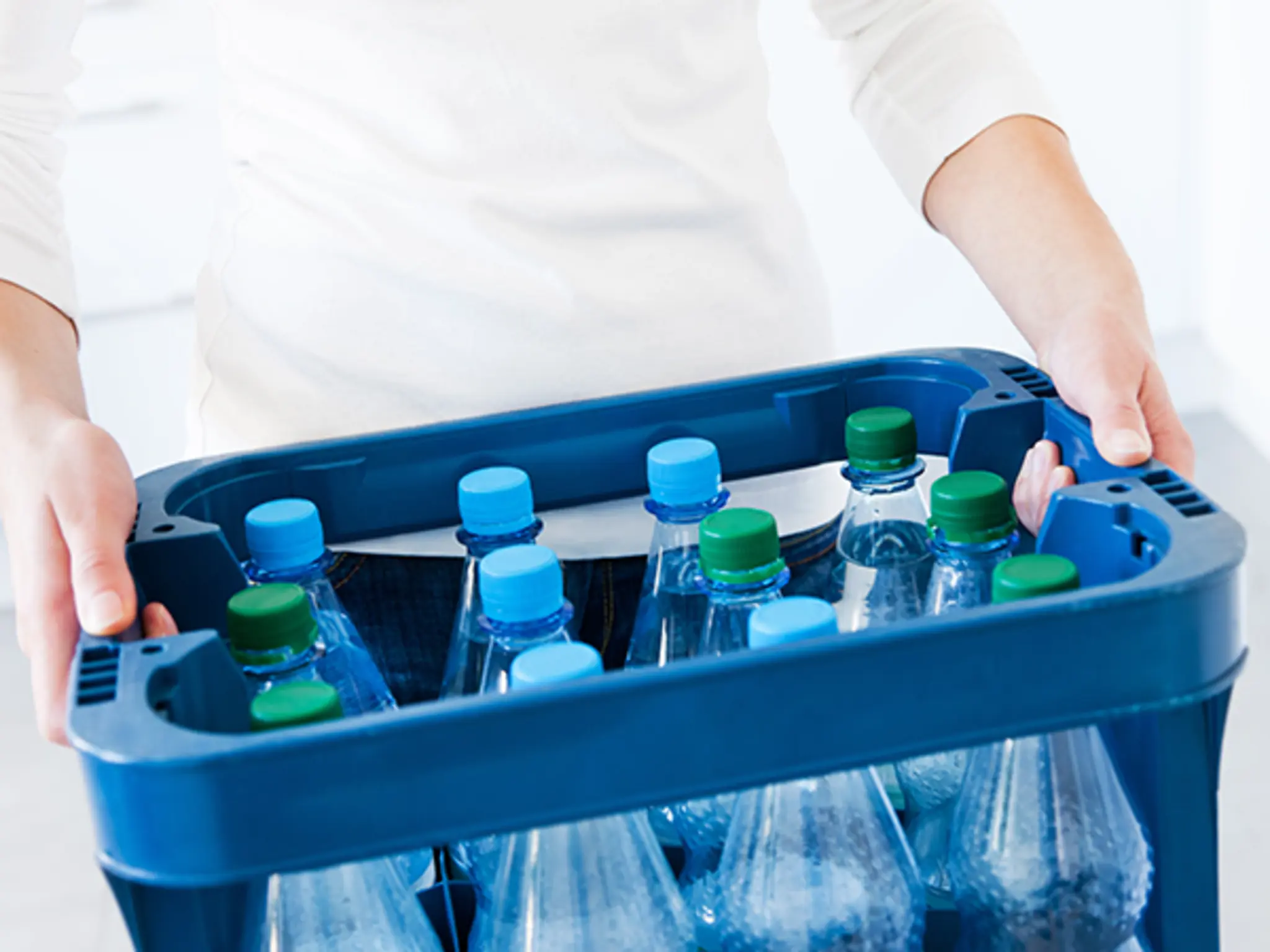Eine Person in einem weißen Oberteil trägt eine blaue Wasserkiste mit Plastikflaschen darin.