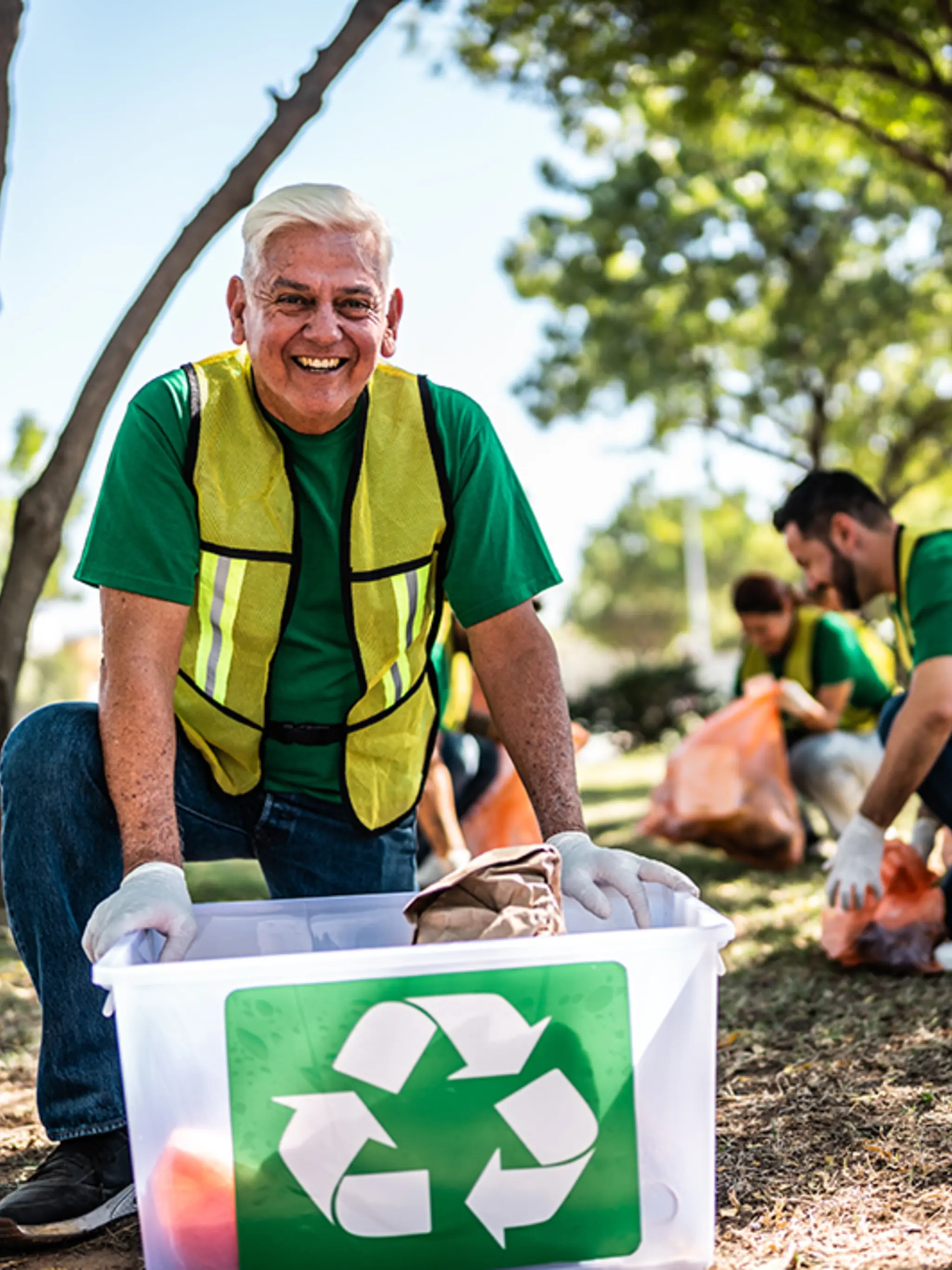 Ein lachender Mann in Arbeitskleidung stützt sich beim Müllsammeln in der Natur auf einer Kiste mit Recyclingsymbol ab