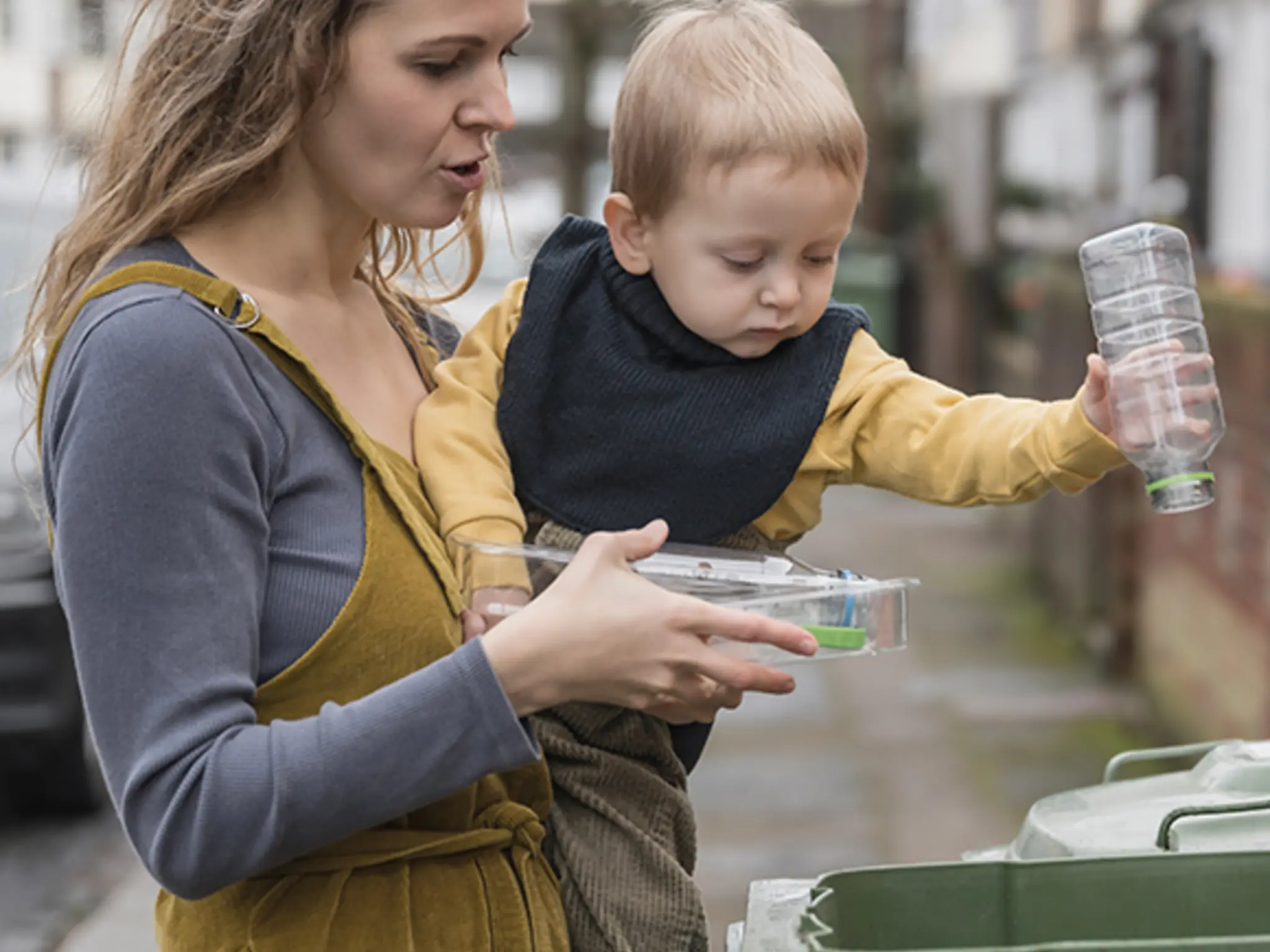 Eine Frau hält ein Kleinkind auf dem Arm und entsorgt mit ihm gemeinsam Plastikflaschen in der Mülltonne 