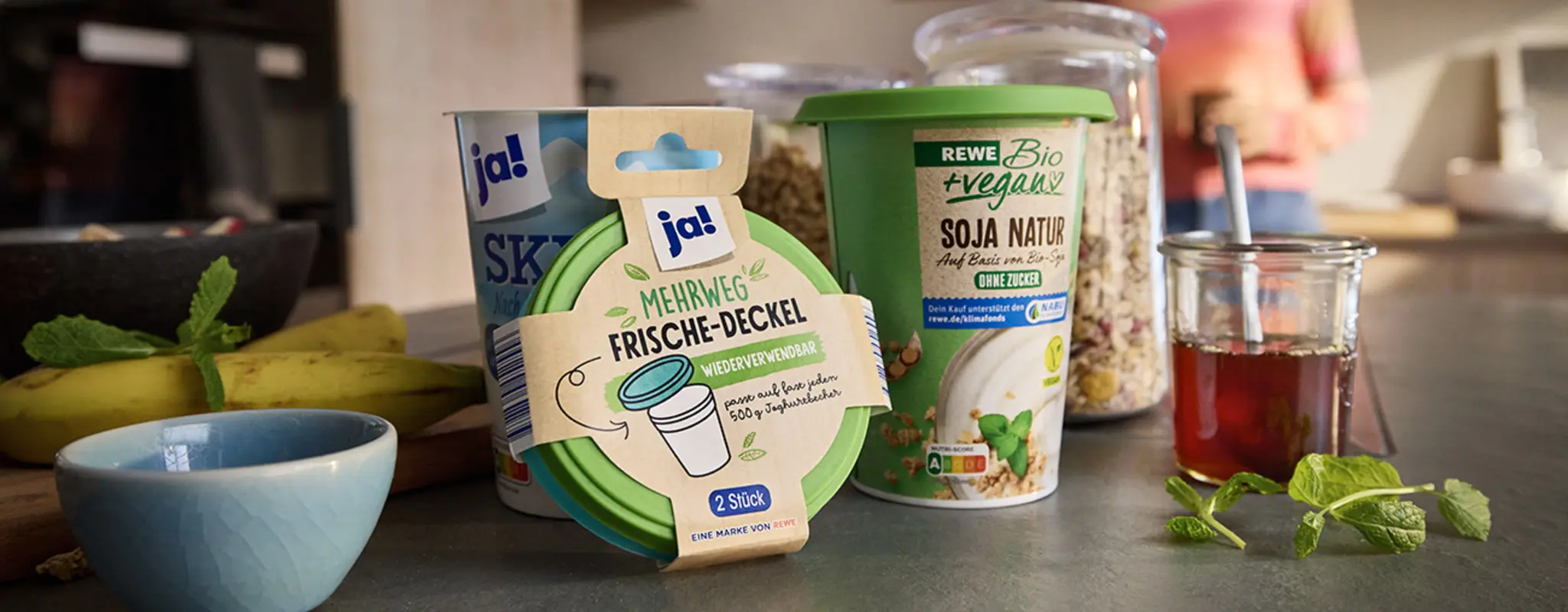 REWE Soja Naturjoghurt und Skyr, eine Schale und ein Mehrweg-Deckel auf einer Arbeitsplatte mit Bananen, Müsli und Tee