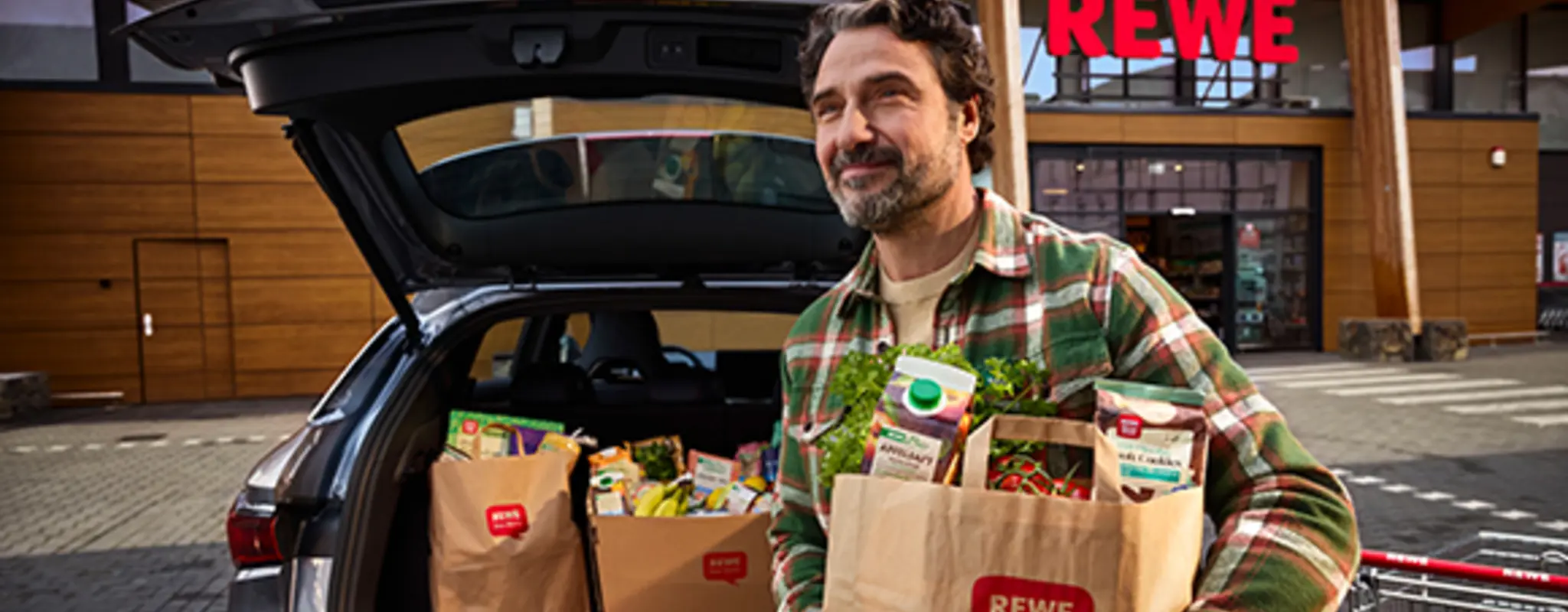 Ein Mann trägt eine mit Einkäufen gefüllte REWE Papiertüte, hinter ihm ein Kofferraum mit weiteren Einkäufen und ein REWE Markt