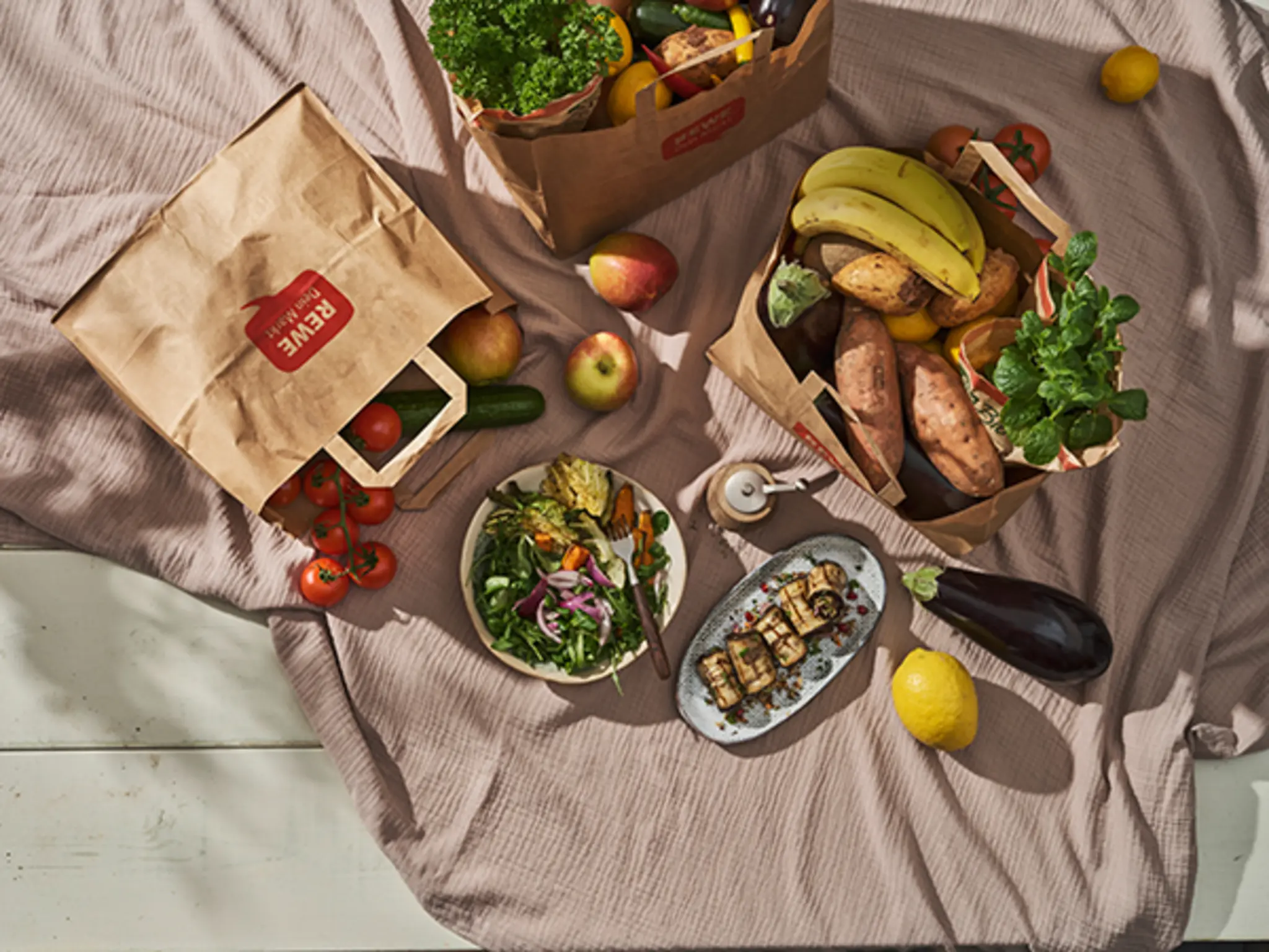 Zwei Teller mit Gemüsegerichten stehen auf einer beigefarbenen Decke, umgeben von REWE-Papiereinkaufstüten gefüllt mit verschiedenem Obst und Gemüse