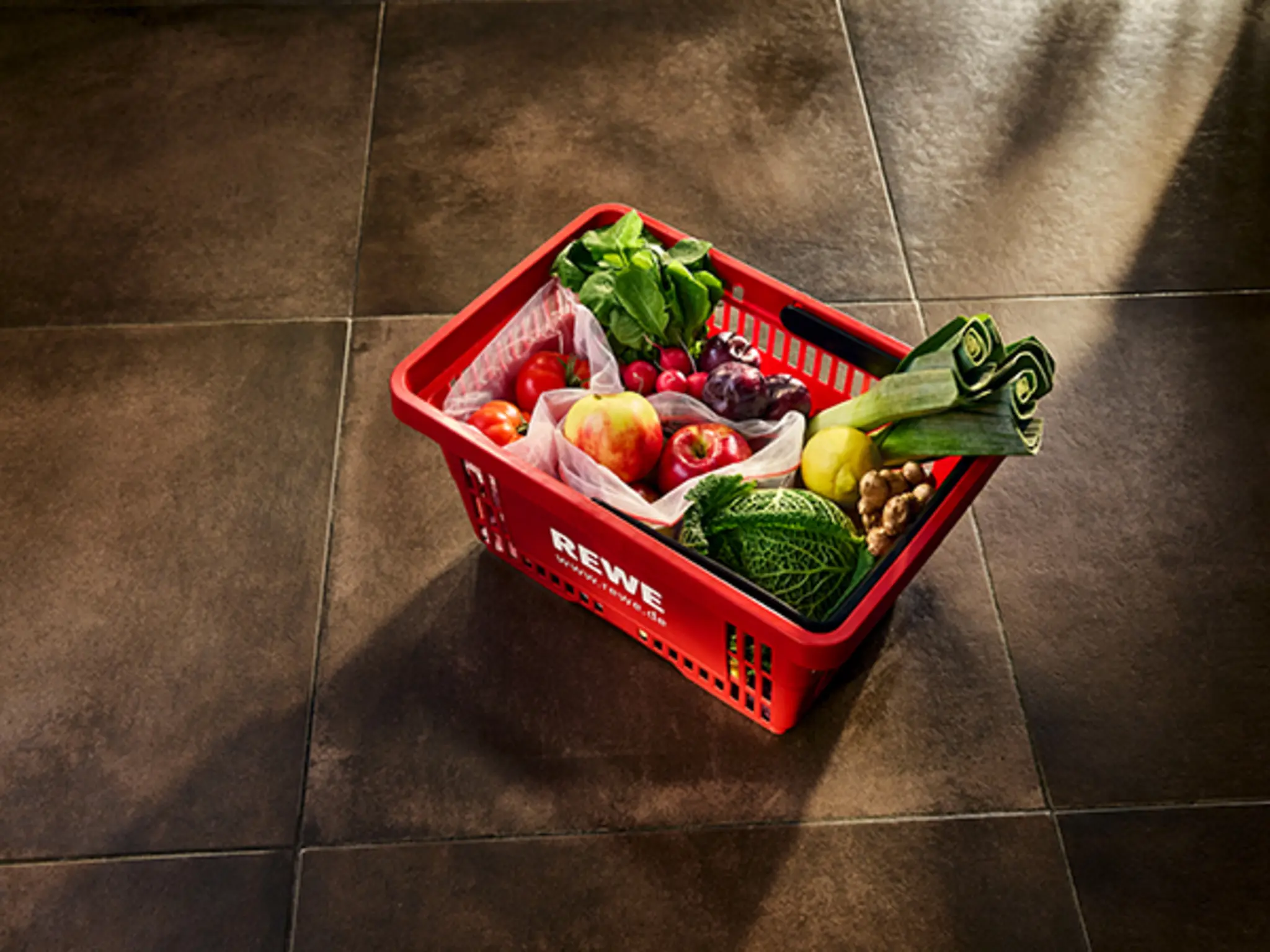Ein REWE-Einkaufkorb auf einem Fliesenboden, gefüllt mit Obst und Gemüse in wiederverwendbaren Netzen