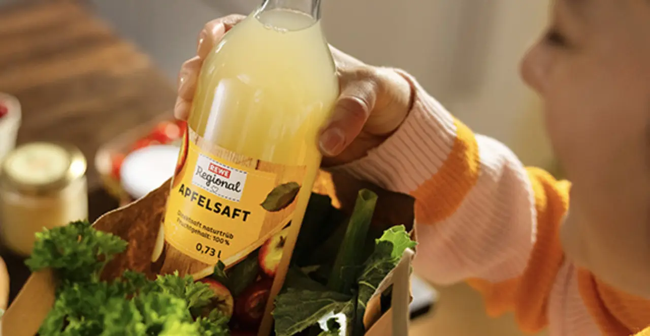 Eine Frau holt eine Flasche REWE Regional Apfelsaft auf einer REWE Papiertragetasche voll frischem Obst und Gemüse.