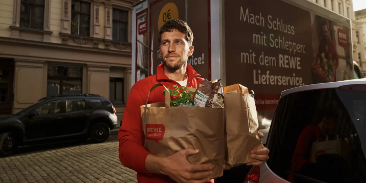 Ein REWE Lieferservice Mitarbeiter trägt Papiertüten mit einem Einkauf.    