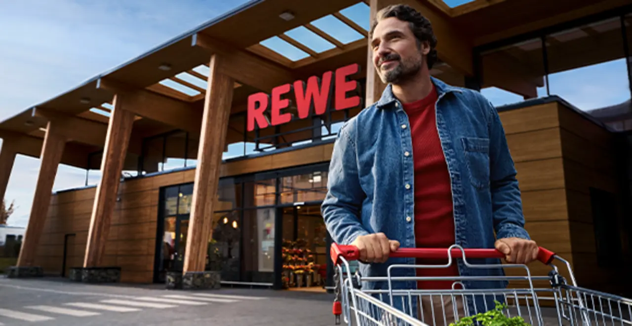 Ein nachhaltigerer REWE Markt mit Holzelementen und Tageslichtarchitektur. Ein Kunde schiebt einen Einkaufswagen über einen weitläufigen Parkplatz. 