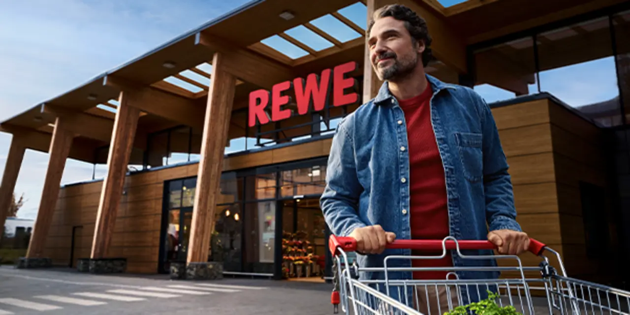 Ein nachhaltigerer REWE Markt mit Holzelementen und Tageslichtarchitektur. Ein Kunde schiebt einen Einkaufswagen über einen weitläufigen Parkplatz. 