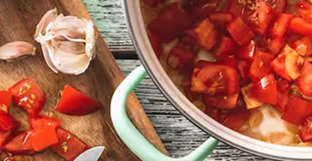 Ein Topf mit gewürfelten Tomaten steht auf einem Tisch. Daneben liegen Tomaten und Knoblauch auf einem Holzbrett.