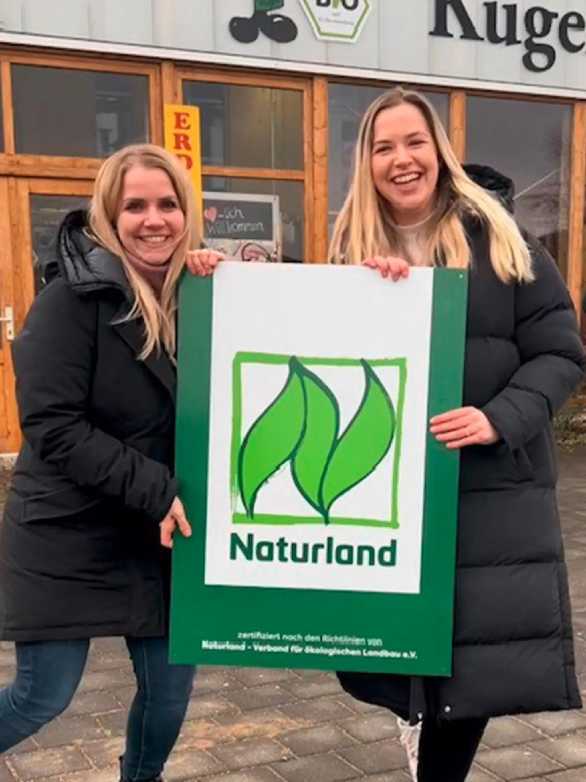 Zwei der Töchter der Familie Kügel halten zwischen sich ein großes Schild mit dem Naturland Zeichen. Als REWE Wegbereiter haben sie sich im Öko-Verband angemeldet.