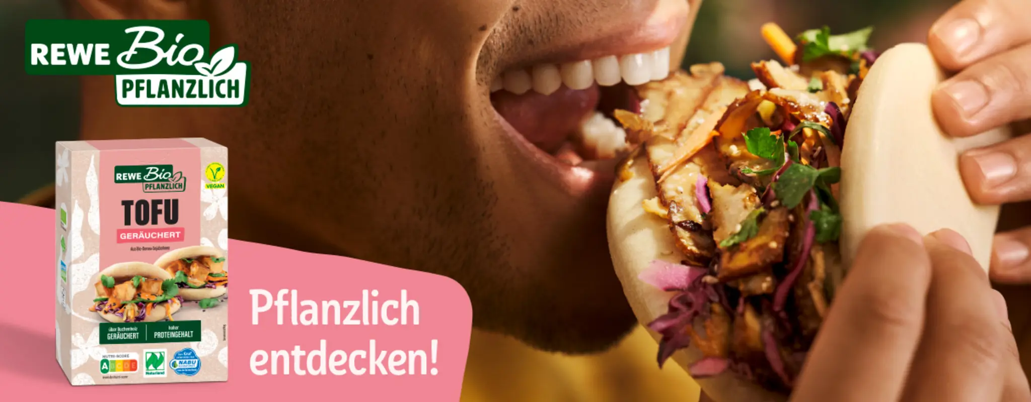 Nahaufnahme einer Person, die in einen pflanzlichen Bao Bun beißt. Auf dem Bild steht das Logo „REWE Bio Pflanzlich“.  