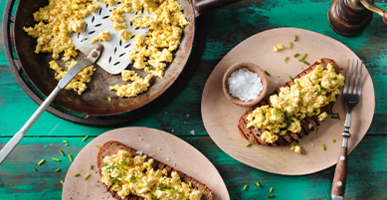 Eine Pfanne Scrambled Tofu steht auf dem Frühstückstisch. Daneben zwei fertige Brote mit Scrambled Tofu.