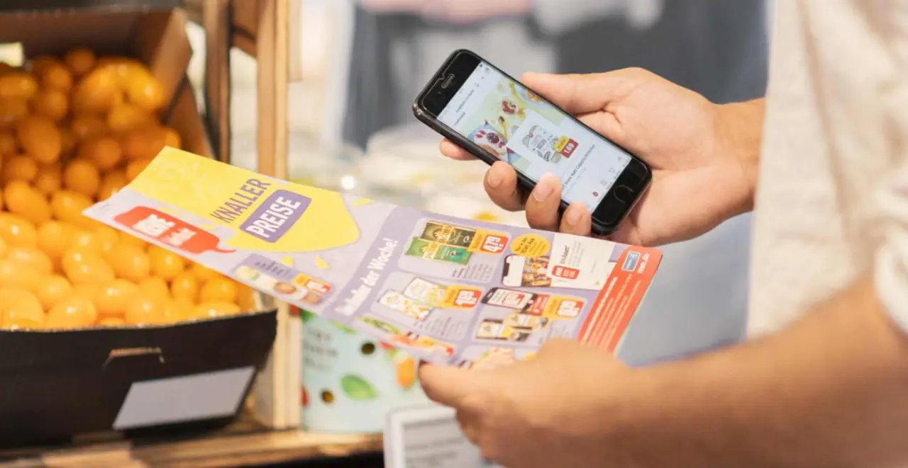 Eine Person hält in der linken Hand einen REWE Papier-Prospekt und in der rechten ein Smartphone mit den aktuellen Angeboten.