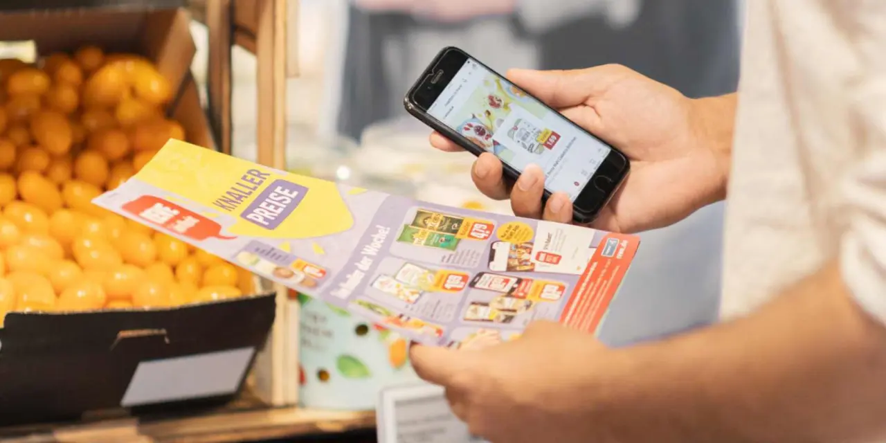 Eine Person hält in der einen Hand den REWE Papierprospekt und in der anderen ein Smartphone.