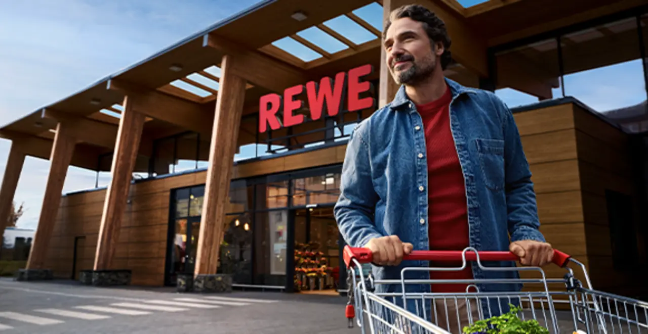 Ein nachhaltigerer REWE Markt mit Holzelementen und Tageslichtarchitektur. Ein Kunde schiebt einen Einkaufswagen über einen weitläufigen Parkplatz.