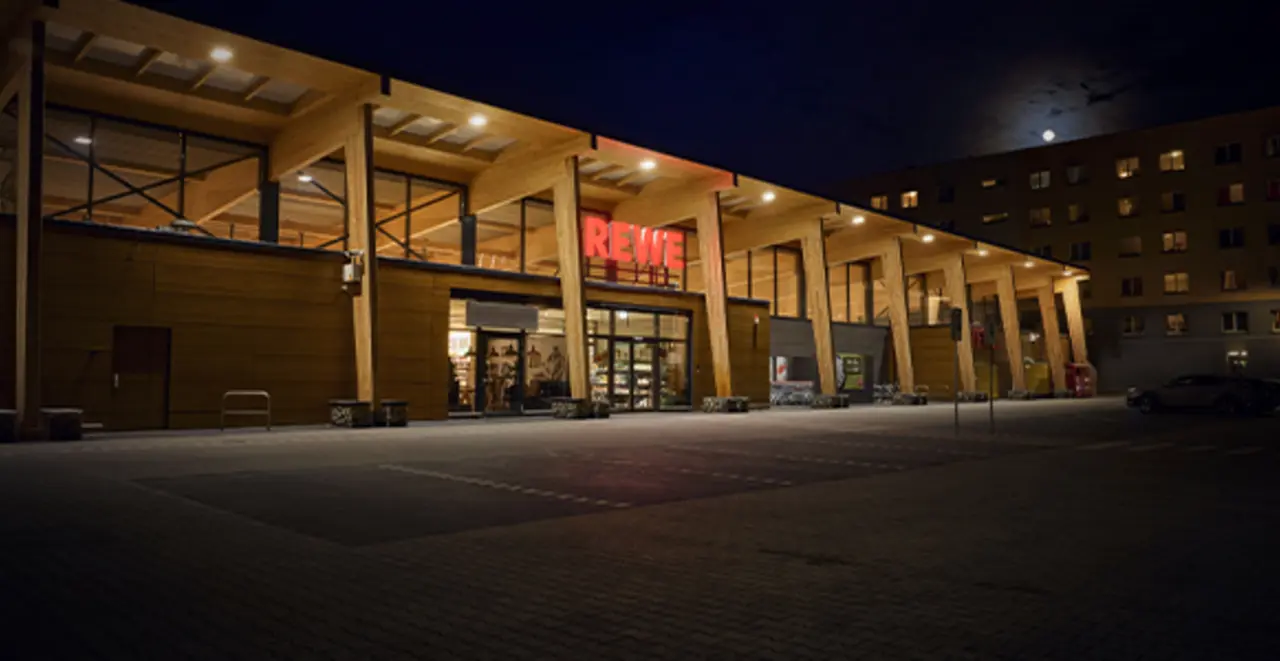 Ein moderner REWE Markt bei Dunkelheit. Licht scheint durch die großzügige Glasfassade. Der Markt wird mit 100 % Grünstrom betrieben.